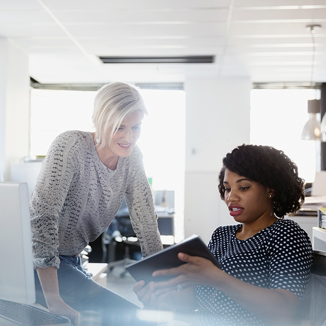 Woman sitting at a desk showing a tablet to another woman who is standing next to her.